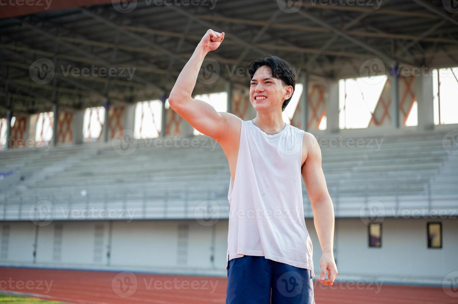 An asian man in white tank top is smiling and raising a fist while standing on racetrack. photo