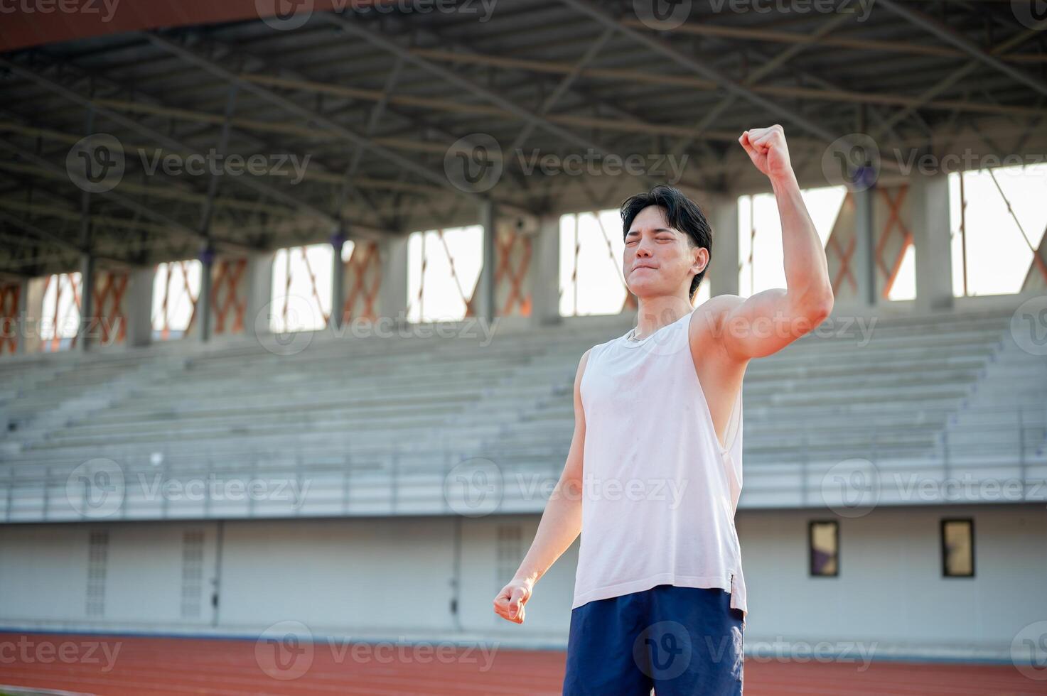 Asian man in white tank top is closing eyes and raising a fist while standing on racetrack. photo