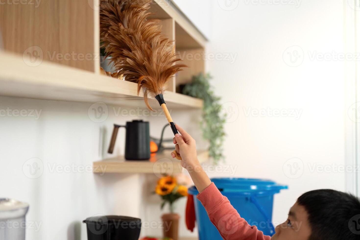 Child Helping with Cleaning. Young boy dusting shelves with a feather duster in a bright kitchen. photo