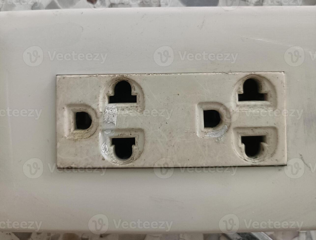 Close up of double electrical outlet with worn and dirty appearance, mounted on white surface, showing signs of age and use, with some dust and minor damage visible photo