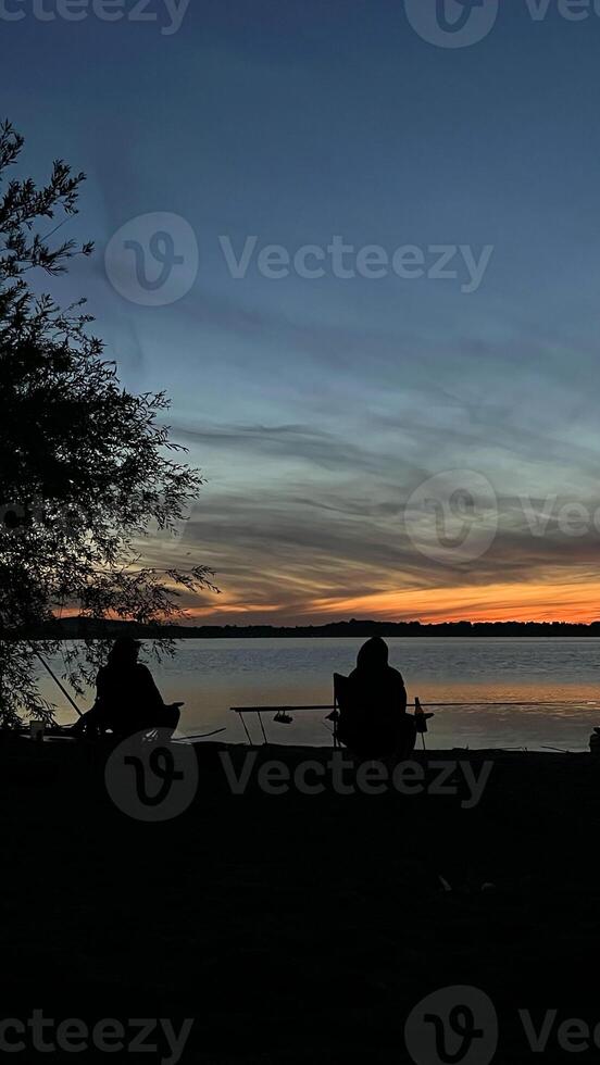 Two fishermen with a fishing rod on the river bank watching the sunset over the water. Poland. photo