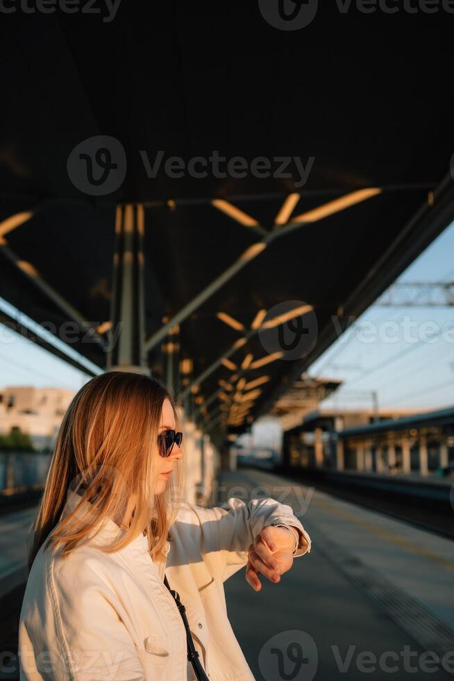 Vertical screen. blonde woman executive wearing sunglasses and white blazer checking wristwatch while waiting near railway tracks during golden sunset lighting. Woman checking time, waiting for train photo