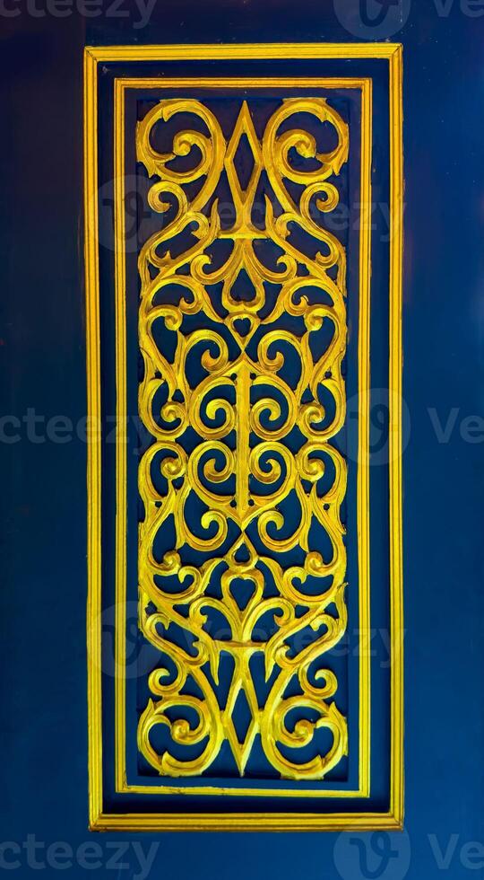 A vibrant close-up of an intricate golden carving, showcasing symmetrical, swirling traditional Thai patterns, set within a rectangular frame against a rich, deep blue background. The golden relief photo