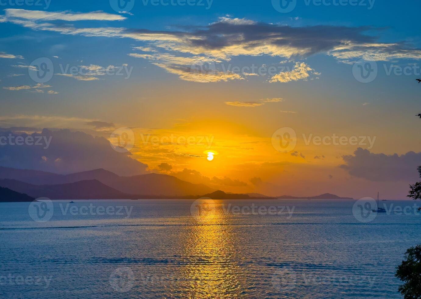 A breathtaking wide-angle shot captures the sun as it dips below the horizon, casting a vibrant golden glow across the tranquil ocean. The distant mountains are silhouetted against the fiery sky photo