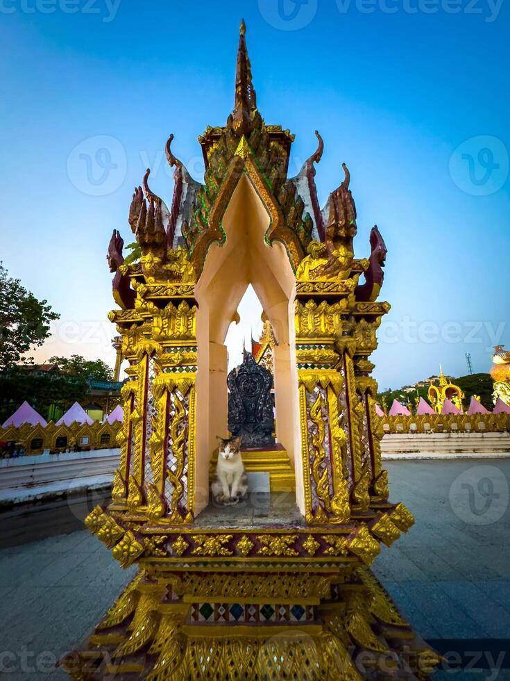 This eye-level, wide-angle shot captures an exquisitely ornate golden shrine, possibly a spirit house or small temple, with intricate detailing and vibrant colors under a clear twilight sky. A white photo
