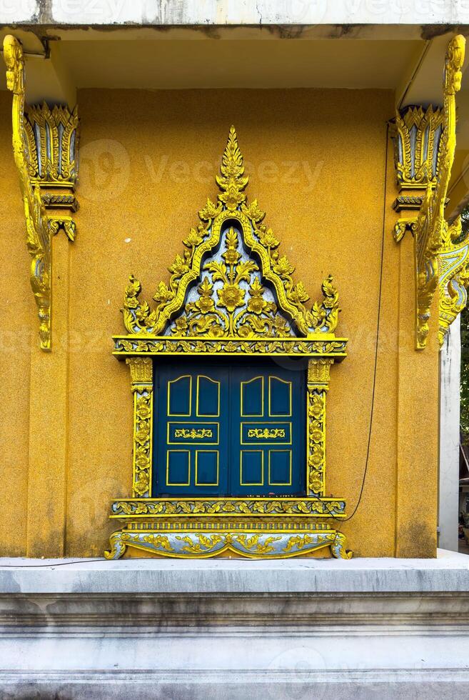 This eye-level, wide-angle shot captures the intricate details of a vibrant golden and blue window, set against a textured ochre wall of a Buddhist temple. The exquisite craftsmanship, typical of photo