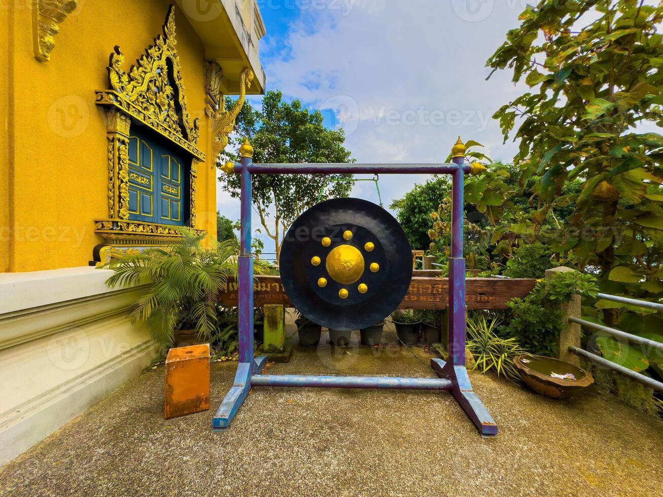 A large, black and gold traditional temple gong hangs from a rustic purple and blue wooden frame, situated on a concrete platform outside a vibrant yellow Thai temple adorned with intricate golden photo