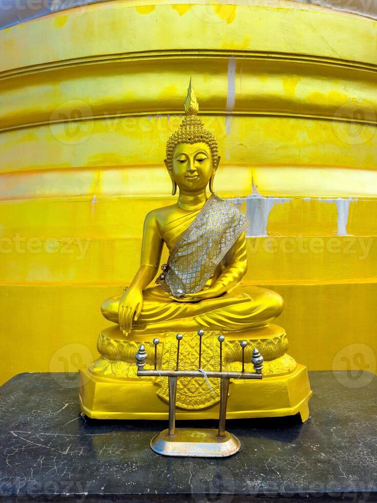 A magnificent golden Buddha statue, adorned with a shimmering silver and gold sash, sits serenely in a meditation pose atop a dark stone surface. The statue is positioned against the vibrant, luminous photo