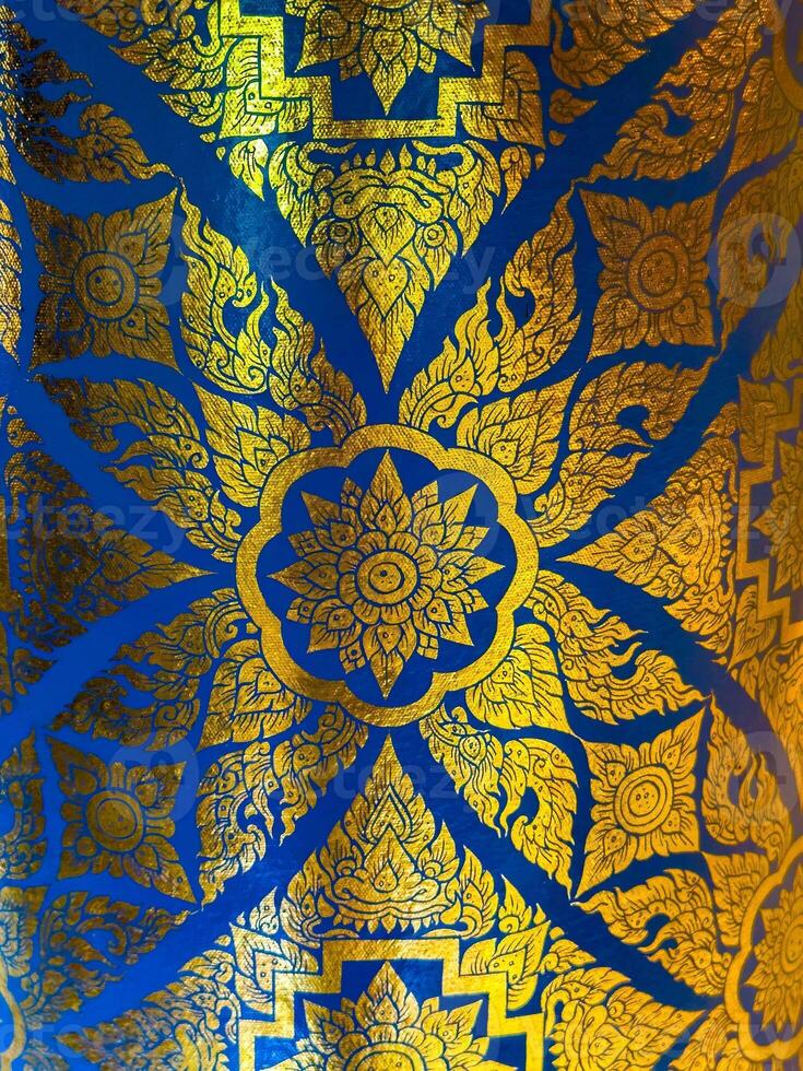 A mesmerizing close-up reveals an ornate traditional Thai pattern, featuring stylized golden floral and geometric motifs, boldly rendered against a rich, deep blue background. The metallic gold photo