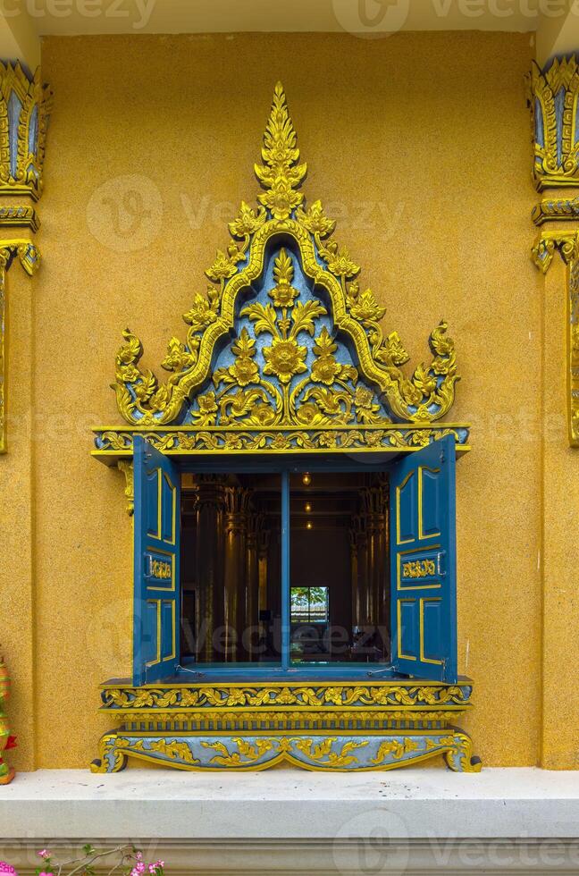 A beautifully detailed window, with its blue wooden shutters opened wide, reveals a glimpse into the richly decorated interior of a Thai temple. The window frame is exquisitely carved with intricate photo