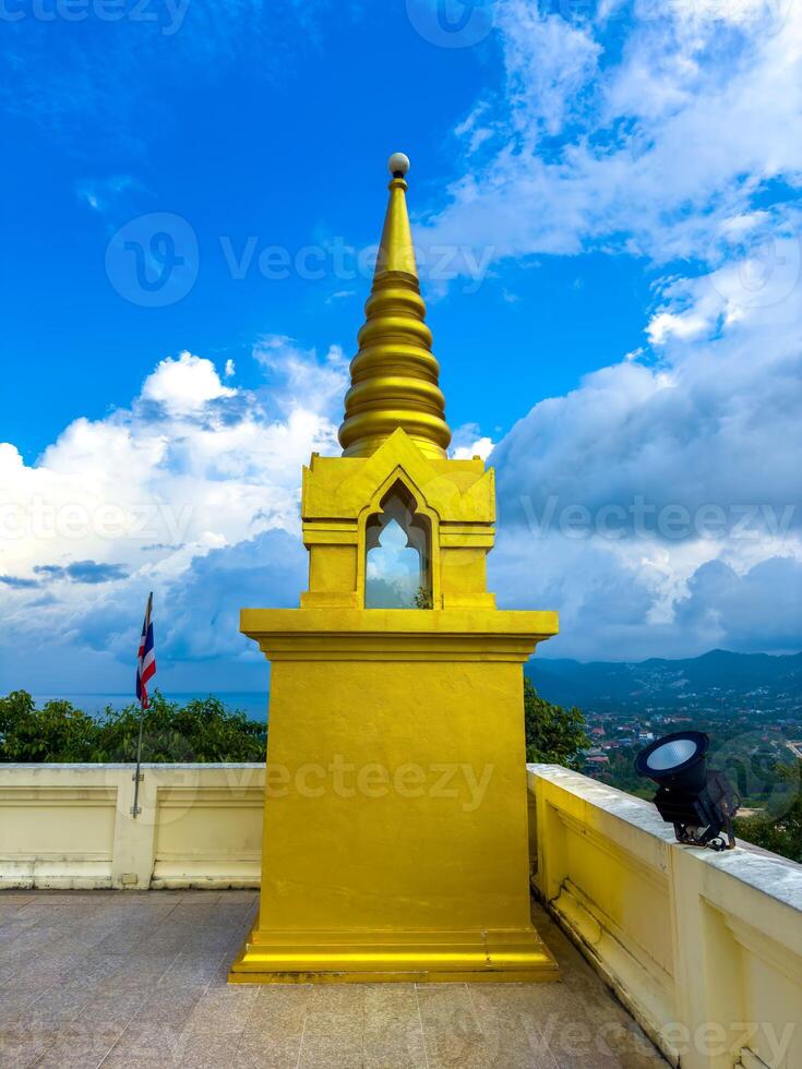 A resplendent golden Buddhist shrine, topped with a tiered spire and featuring a small mirrored window, stands prominently on a white stone terrace. Behind it, a Thai flag flutters gently, and lush photo