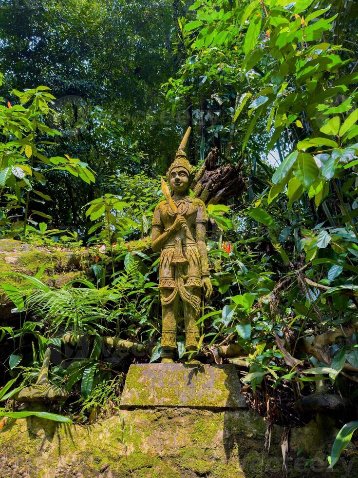A low-angle, medium shot captures a weathered, moss-covered stone statue of a traditional Thai deity standing majestically amidst the dense, sunlit tropical jungle on Koh Samui, Thailand. The photo
