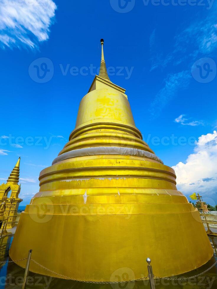 A towering, shimmering golden stupa, with its distinct tiered base and sharp spire, dominates the frame, captured from a low-angle perspective looking upwards. The brilliant gold surface reflects the photo
