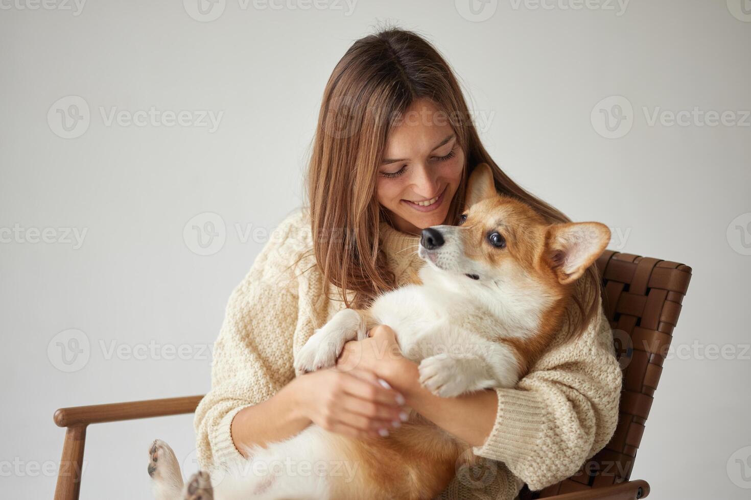 Happy woman holding adorable Corgi dog in cozy sweater, bonding moment on neutral background. photo