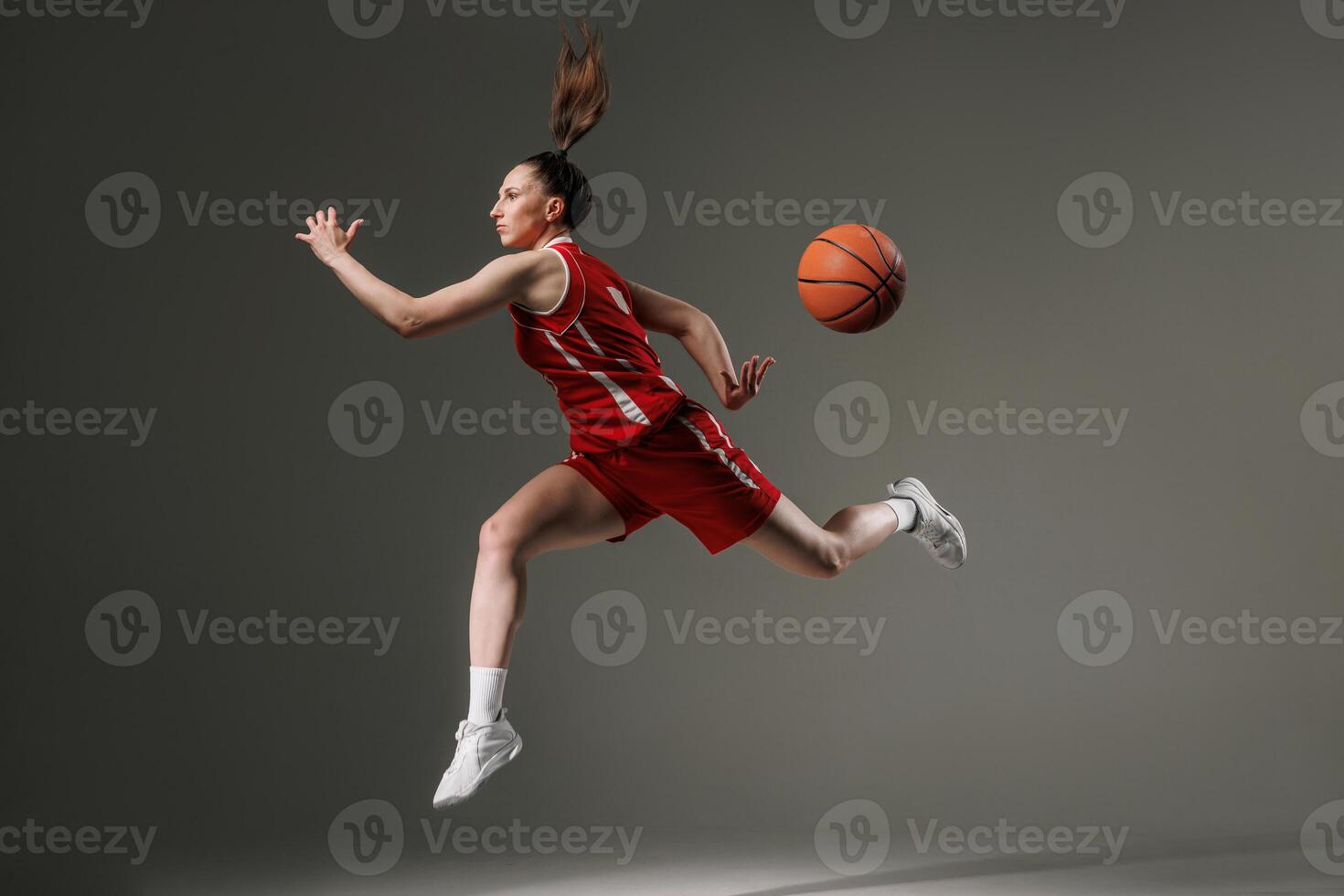 Dynamic female basketball player in mid-air jump with flying ball, wearing red uniform on gray studio background symbolizing strength, motion, and athletic performance. photo