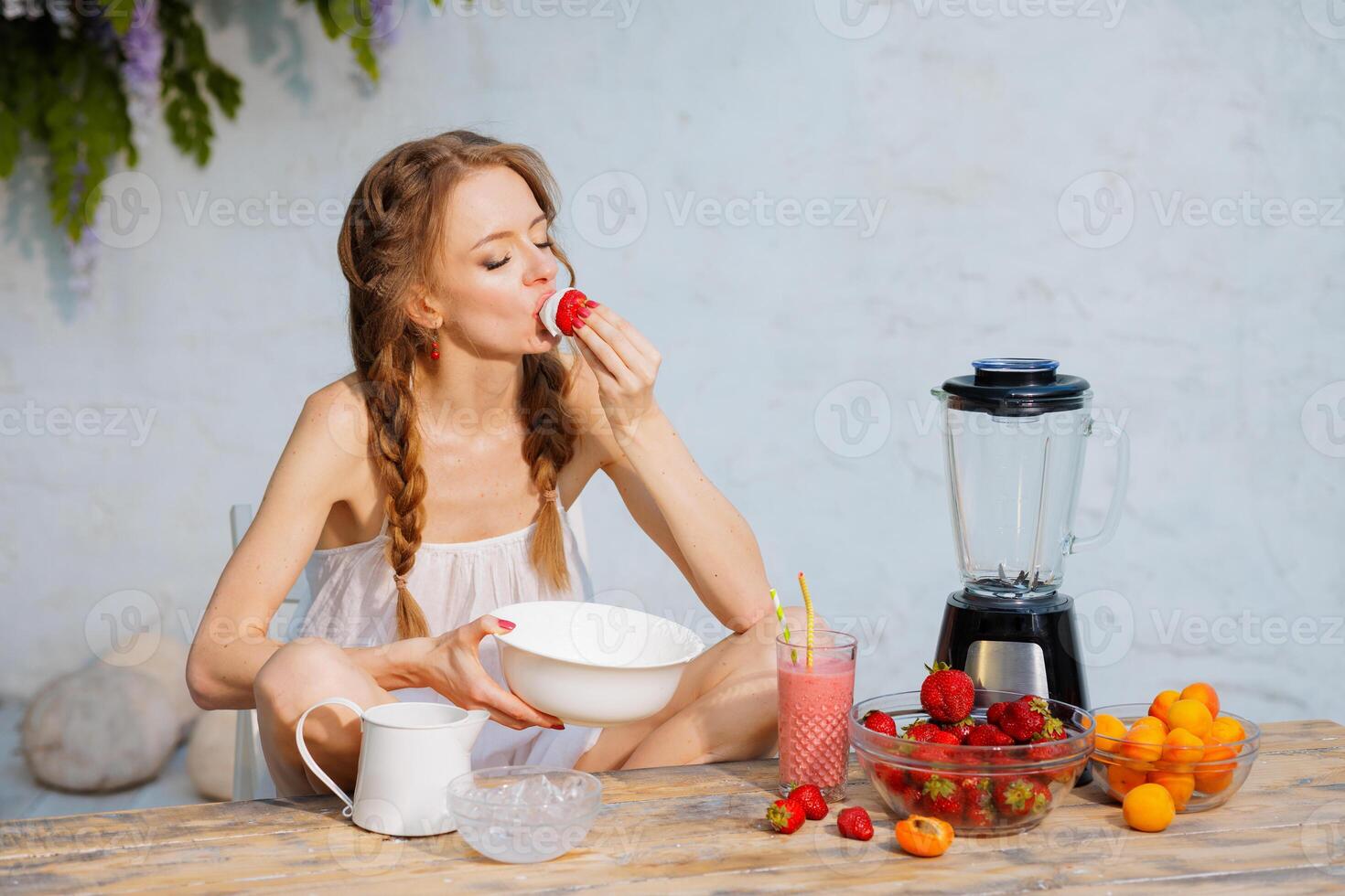 Young woman enjoying strawberries with whipped cream outdoors on a summer day, sitting at rustic wooden table with fresh fruit, smoothie, and blender. Natural healthy dessert concept. photo