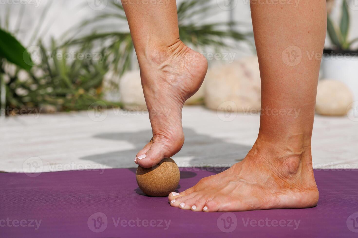 Close up of womans foot massaging on cork therapy ball for plantar fasciitis relief, recovery, and reflexology on a yoga mat outdoors. Wellness, self-care, health, and fitness concept. photo