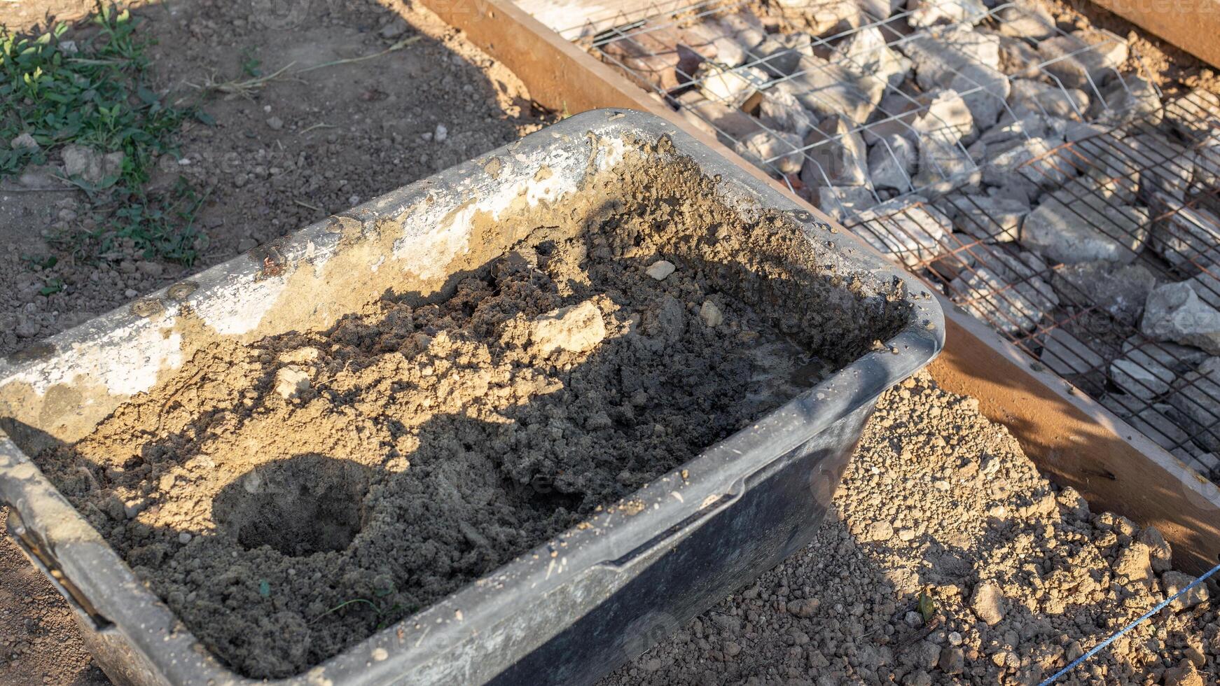 A black tub filled with a mixture of sand and water sits next to a construction site prepared with rocks and wire mesh for pouring concrete photo