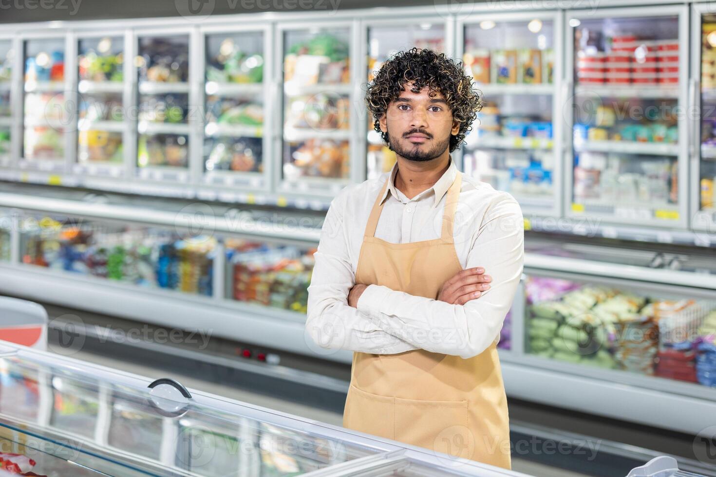 A supermarket worker, wearing an apron, stands in front of the refrigerated section with arms crossed. photo