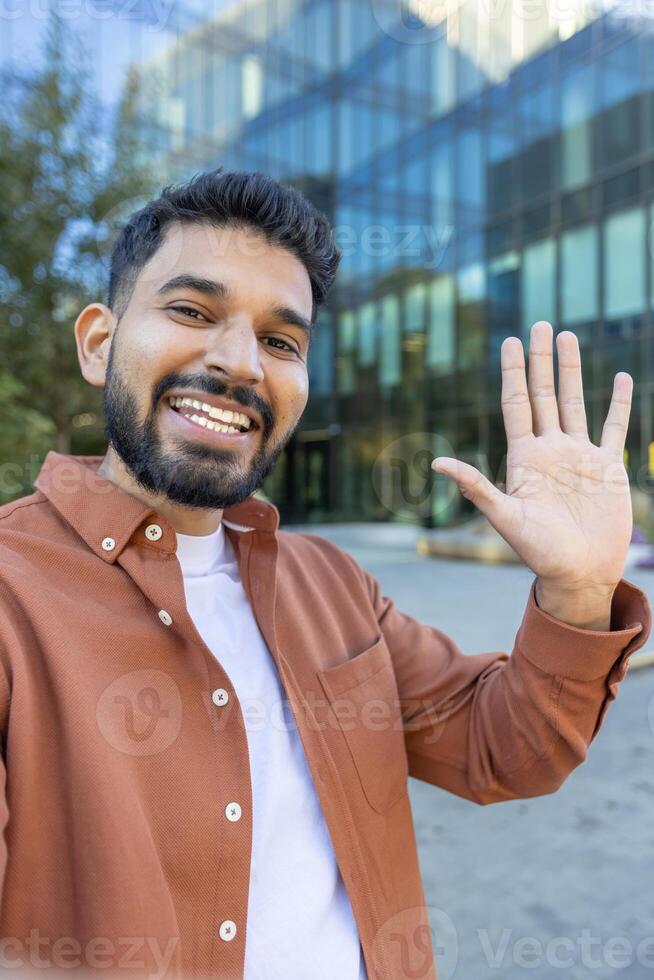 A smiling man on a call outside a modern building, waving at the camera photo
