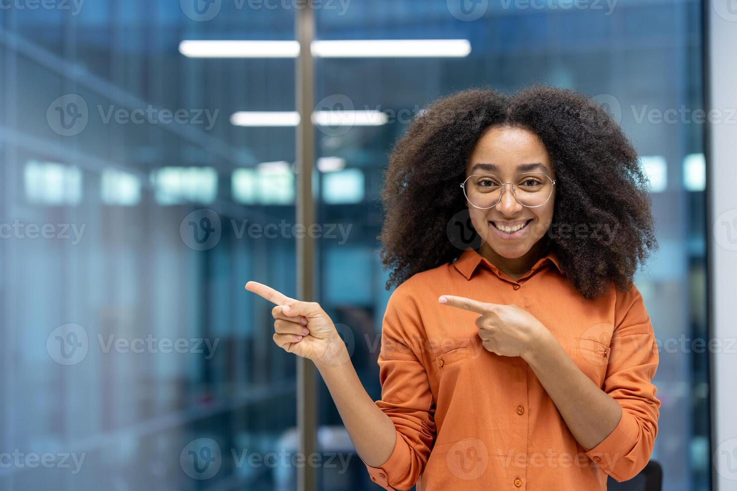 Smiling young woman with curly hair points towards something, framed by a windowed office backdrop. photo