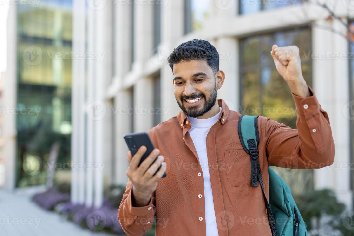 An excited young man looks at his phone while celebrating good news outdoors, raising his fist in triumph. photo