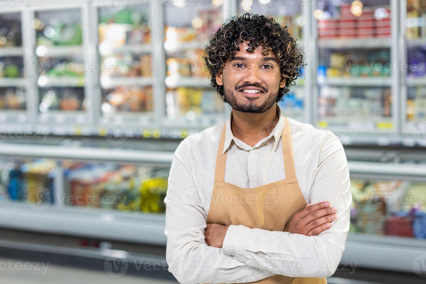A friendly grocery store employee wearing an apron stands in a supermarket aisle with arms crossed, smiling at the camera. photo