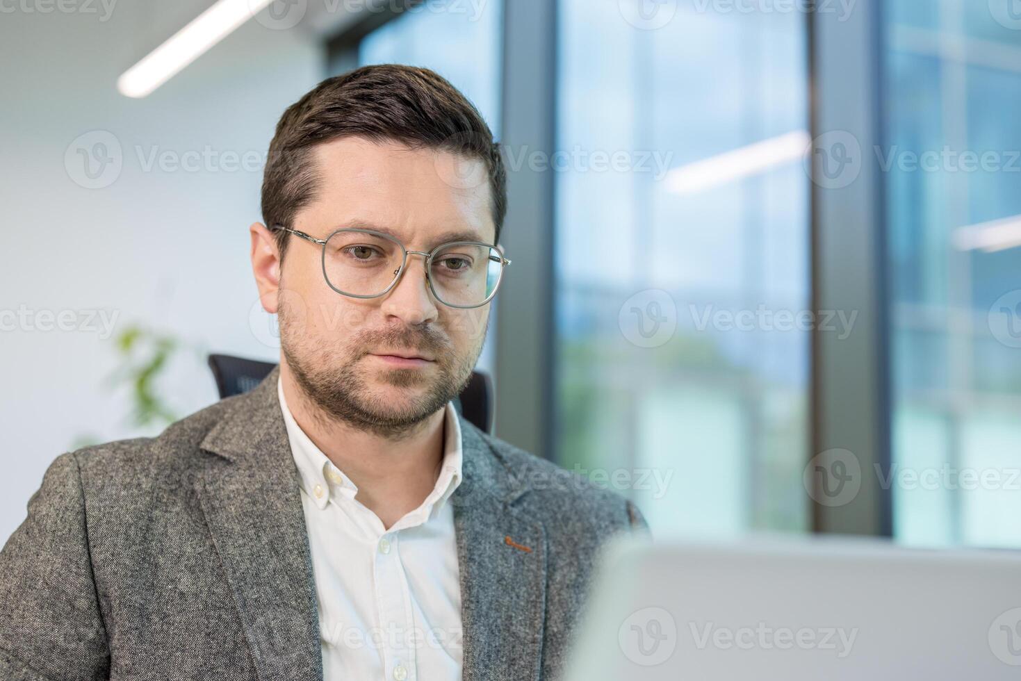 A close-up photo of a young man working in an office at a laptop, looking at the screen with concentration and thoughtfulness.