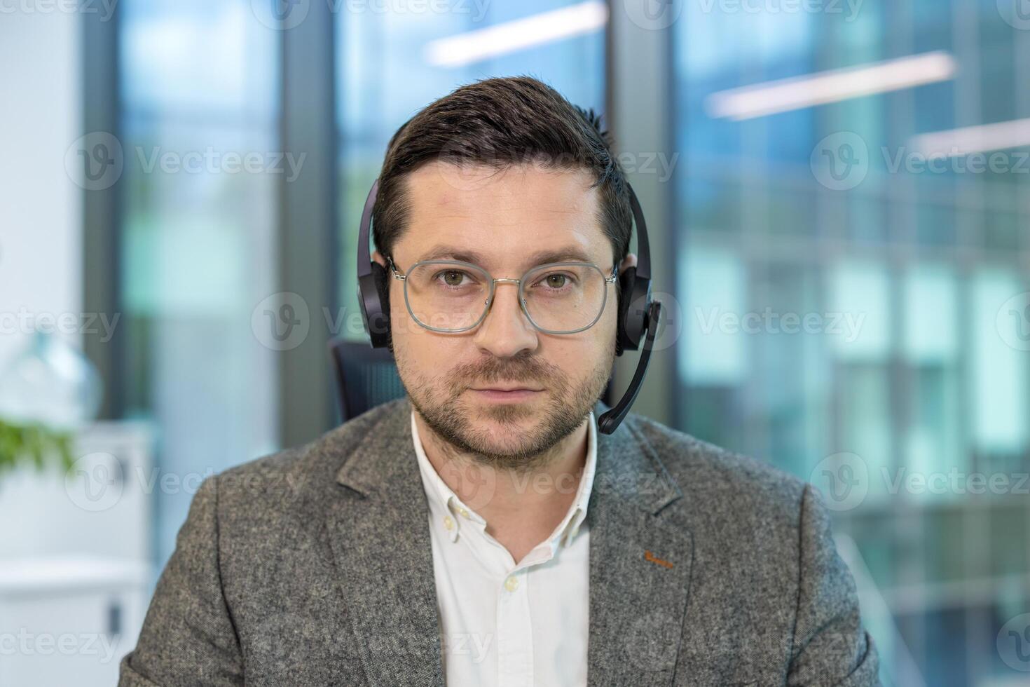 Close-up portrait of a young serious male office worker and assistant sitting at his workplace in a headset and looking intently at the camera. photo