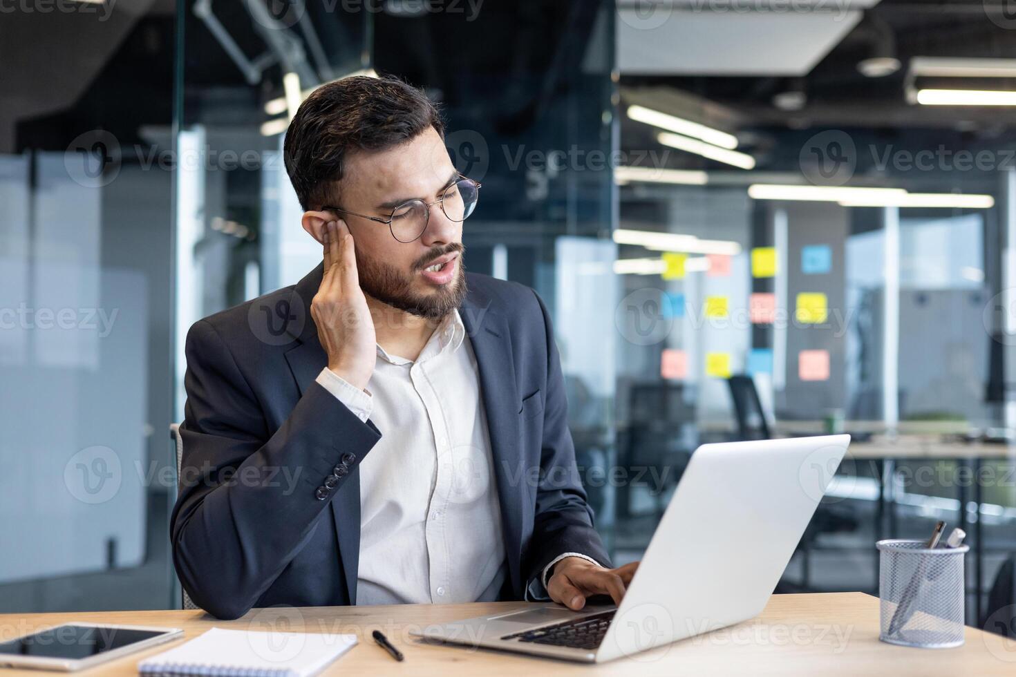 A man in a suit, experiencing ear pain while working on a laptop in an office setting photo