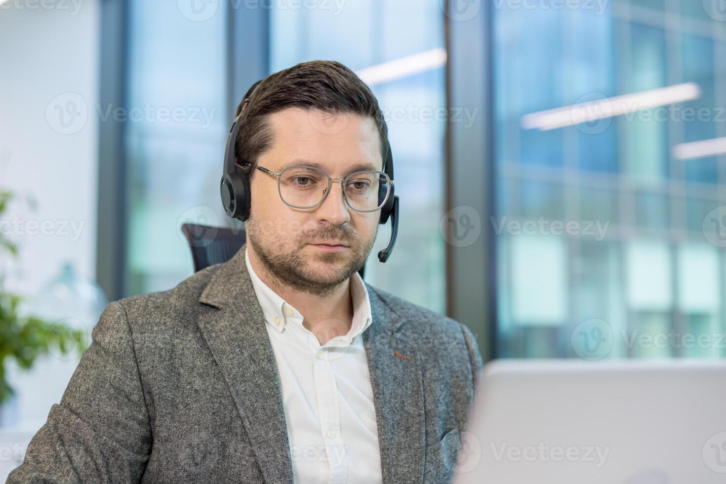 Close-up photo of a young man in a suit and headset working seriously in the office on a laptop.