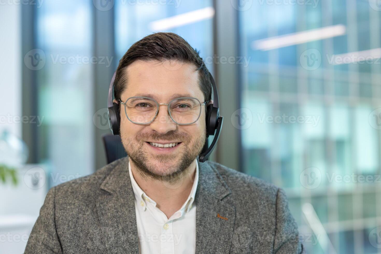 Close-up portrait of a young male office worker sitting at his workplace in a headset and suit and looking at the camera with a smile. photo