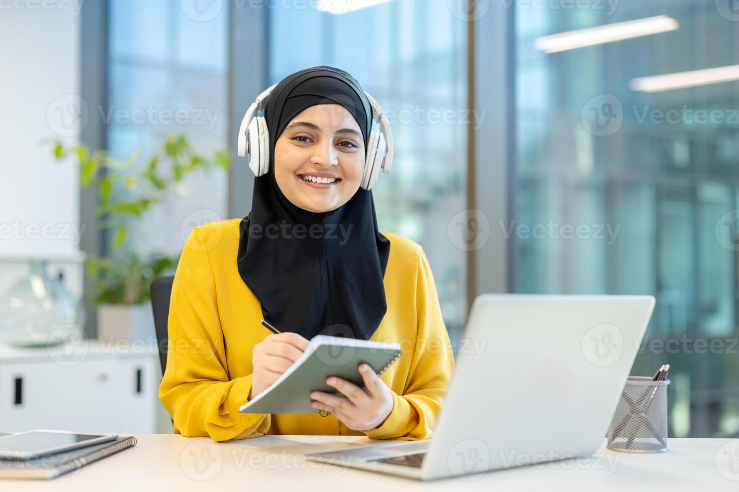 A smiling Muslim woman wearing a hijab and headphones, working on a laptop and taking notes at her desk in a modern office. photo