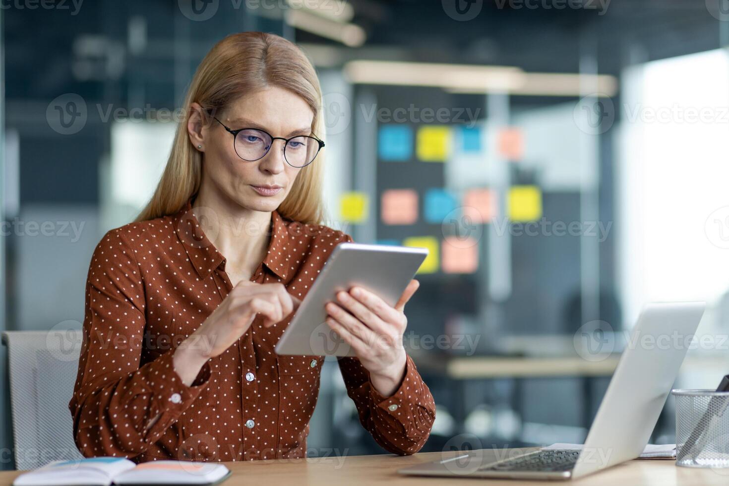 A businesswoman is working at her desk with a tablet and laptop in a modern office setting, wearing glasses and a button-down shirt. photo