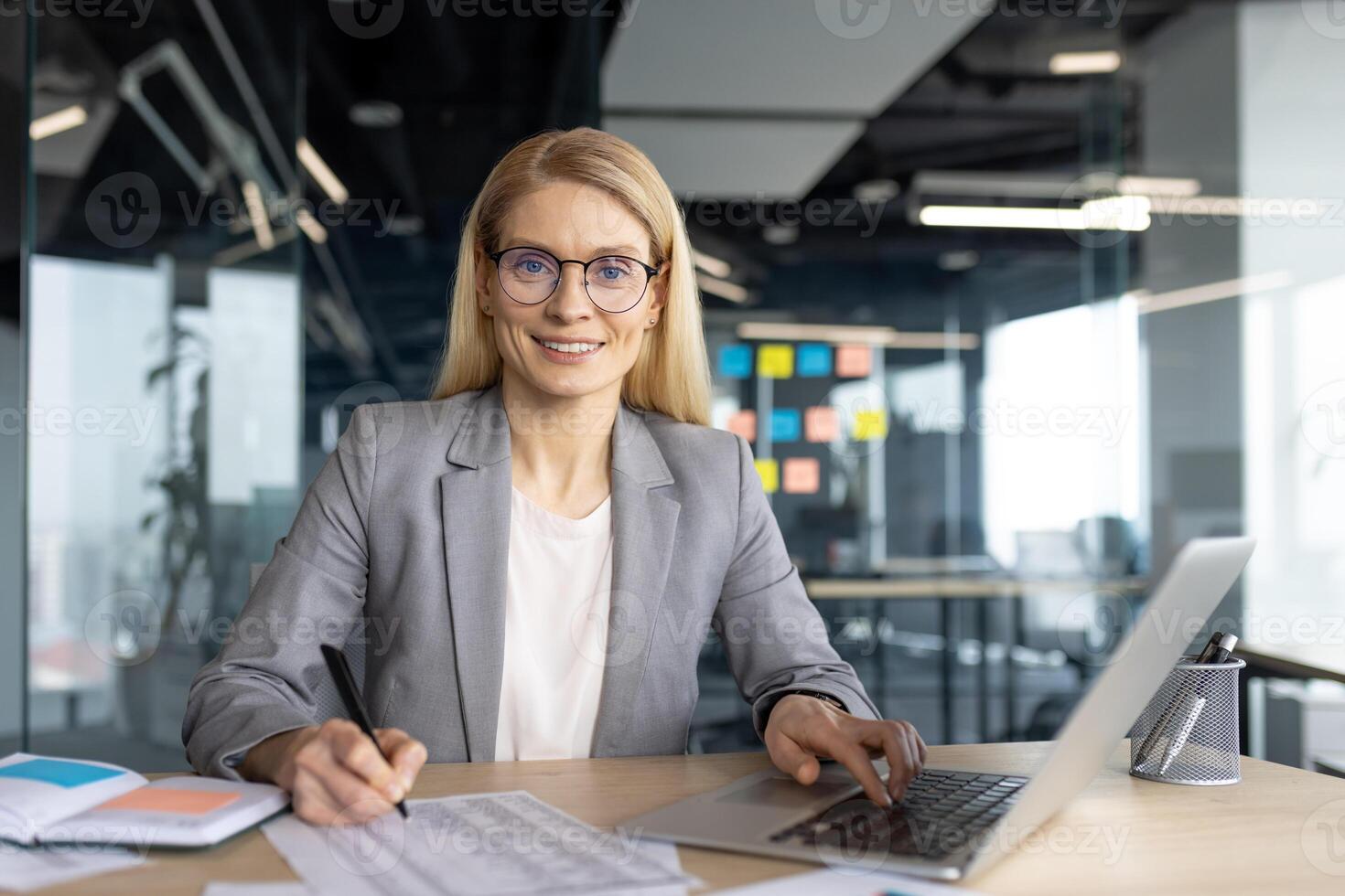A smiling businesswoman in a modern office setting, working on a laptop while taking notes, radiating confidence. photo