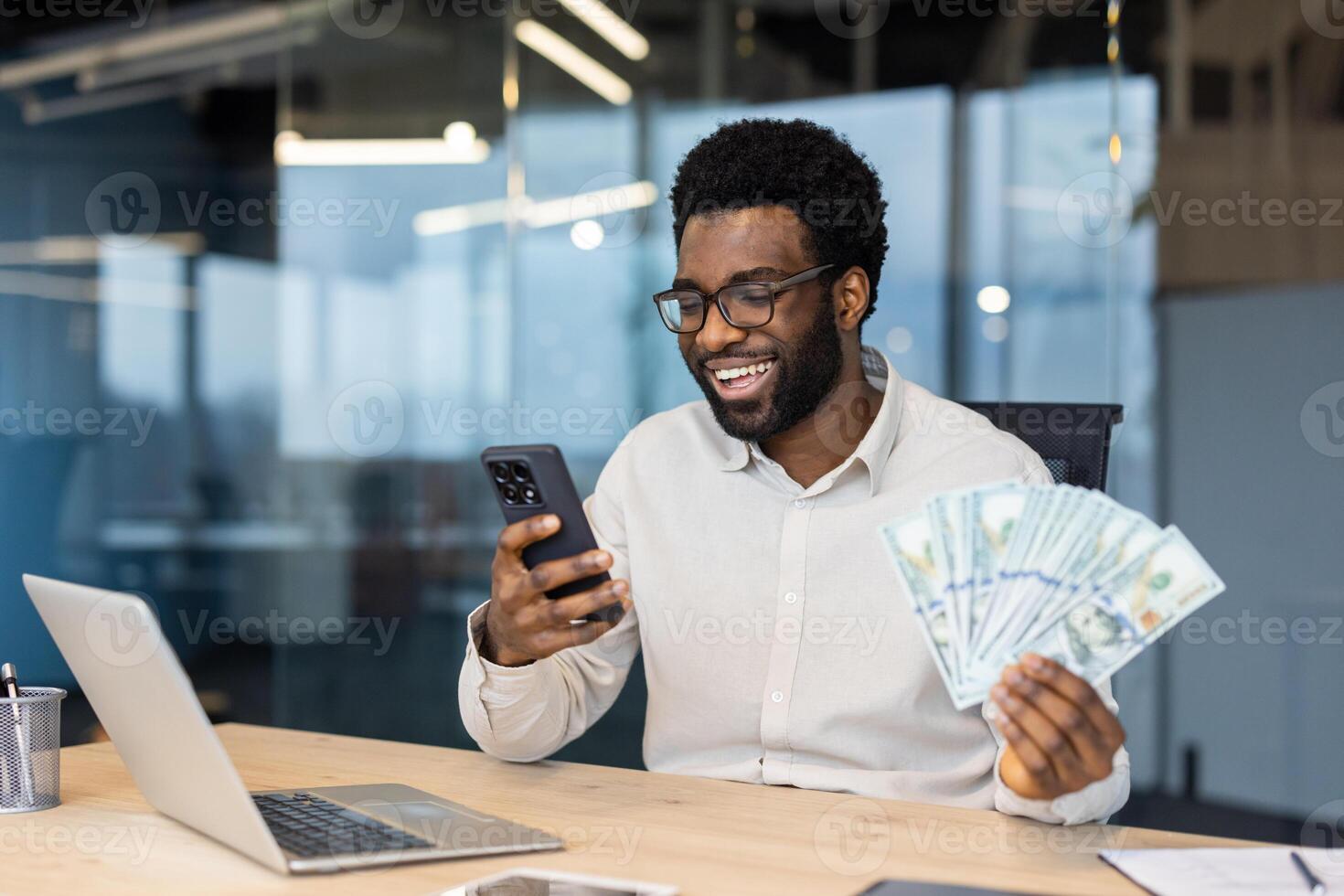A smiling Black man in glasses holding money and looking at his phone, likely at good news in an office setting. photo