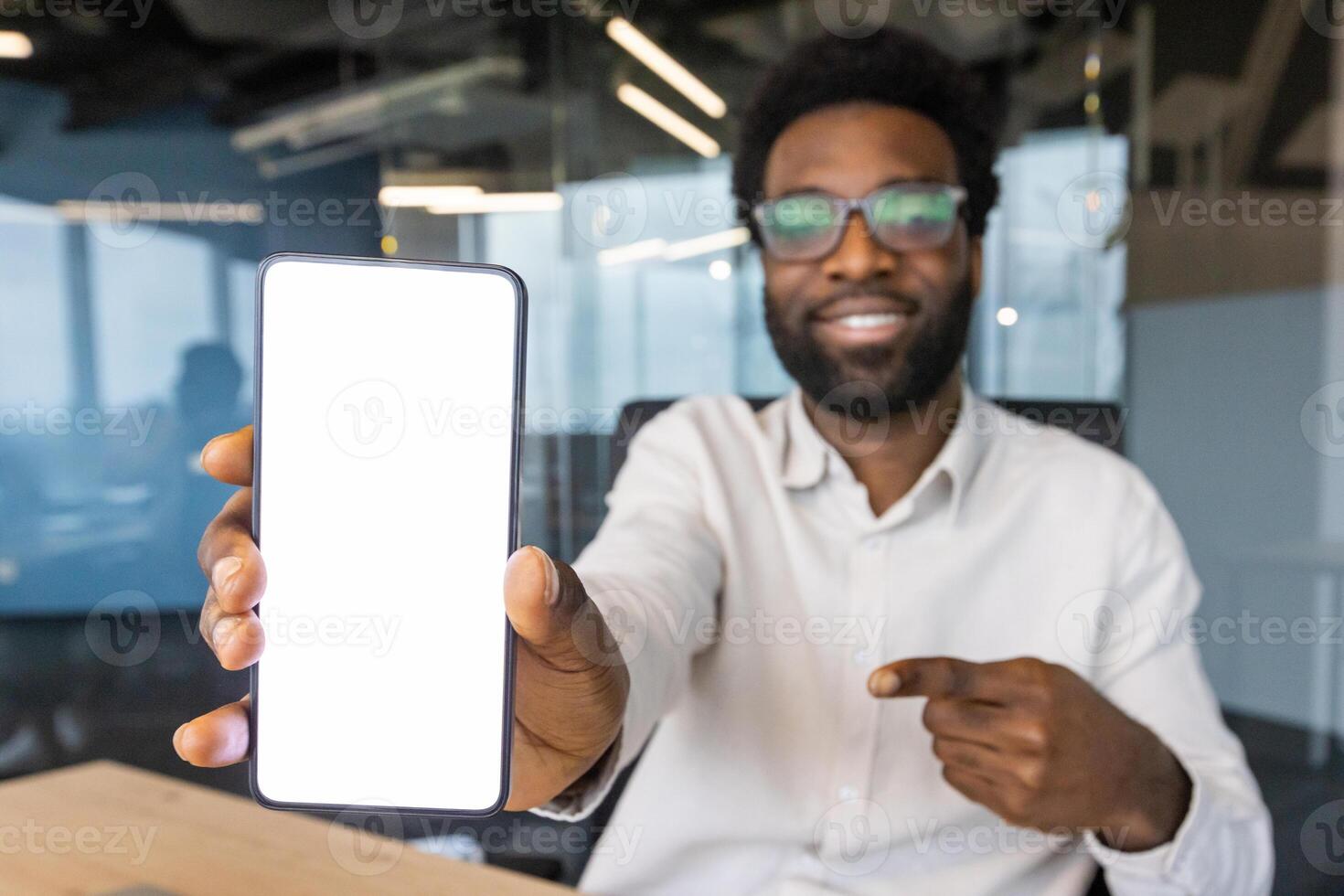 An African American man in glasses points to a blank smartphone screen, smiling confidently at the camera in an office setting. photo
