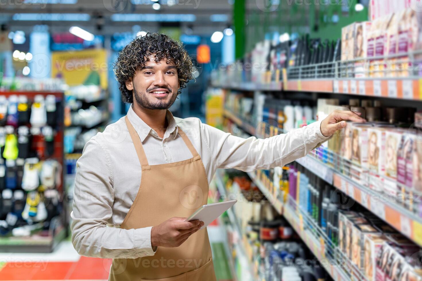 A smiling worker in a supermarket stands by shelves, checking inventory with a tablet while wearing an apron. photo