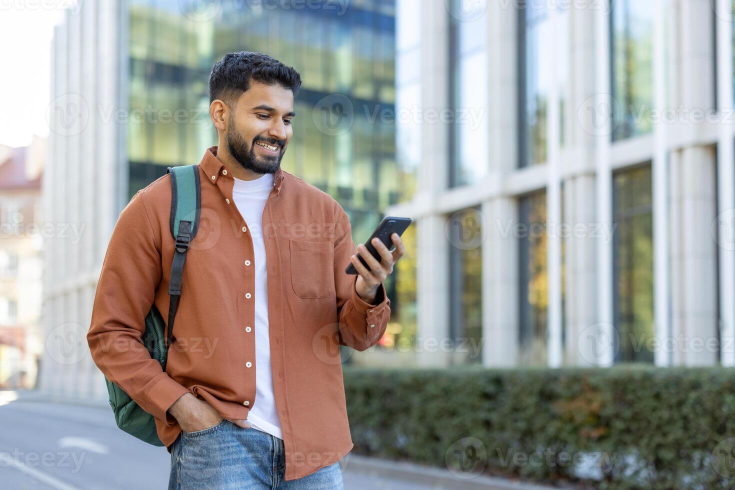 A smiling student, outdoors, using his phone while walking in front of a modern building photo