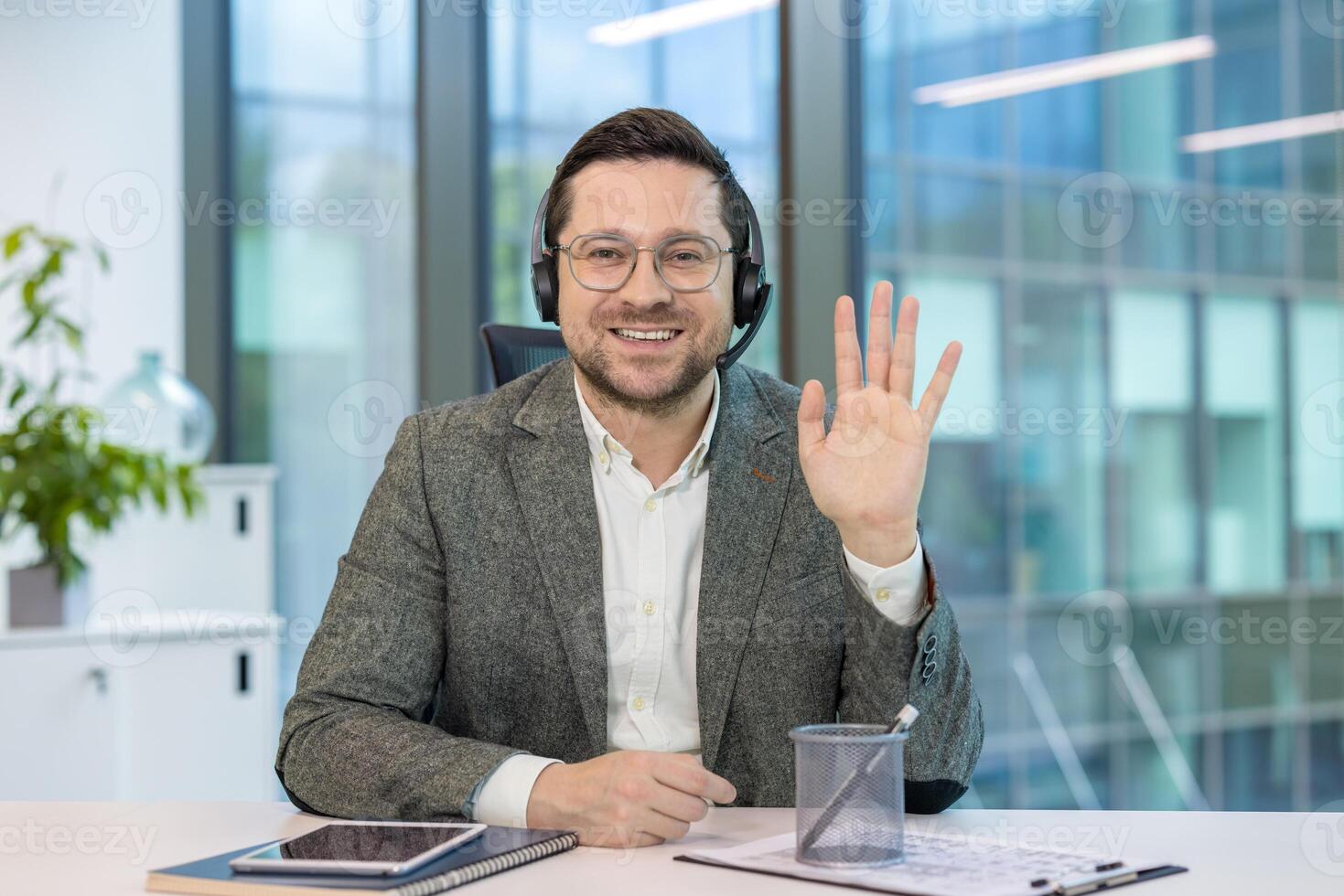 Portrait of a businessman in a headset and suit sitting at a desk in the office, smiling and waving at the camera. photo