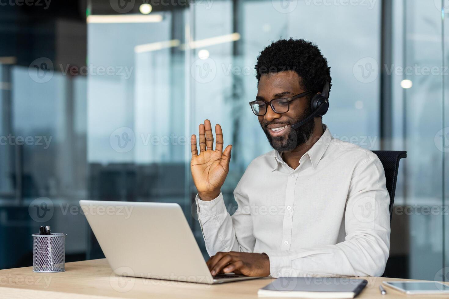 A smiling man in a call, wearing a headset and glasses, waving at a laptop in an office setting. photo