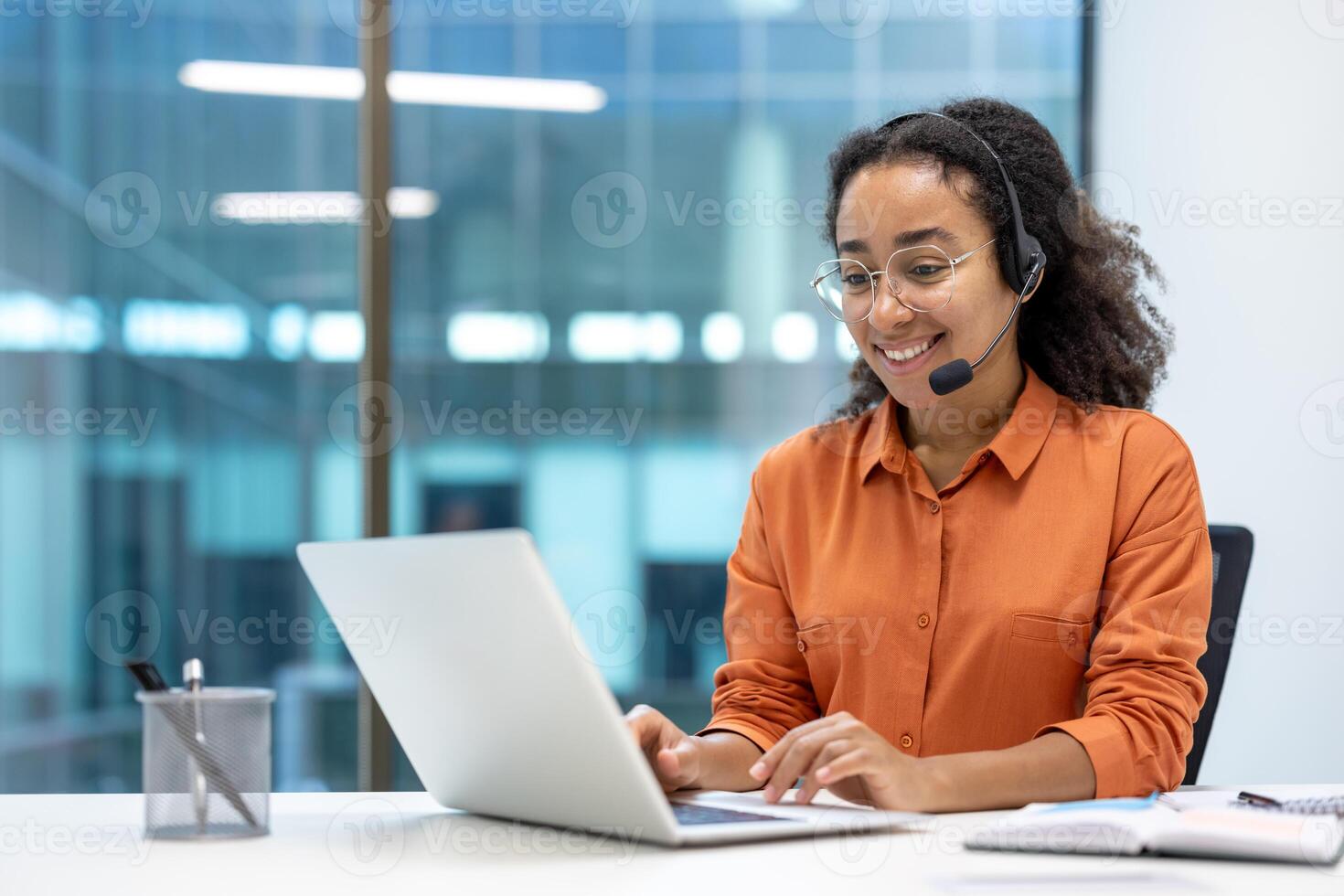 Smiling customer service representative wearing a headset and glasses working on a laptop in a modern office setting. Focus on communication and support. photo