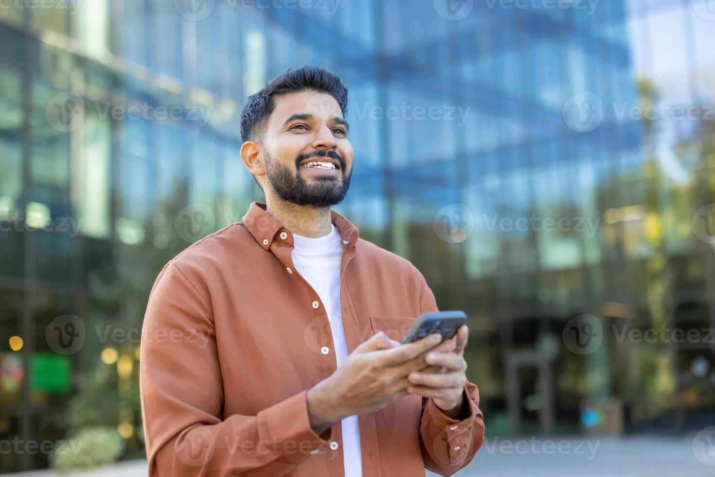 A smiling man holds a smartphone in front of a modern glass building, suggesting a business or technology context. photo