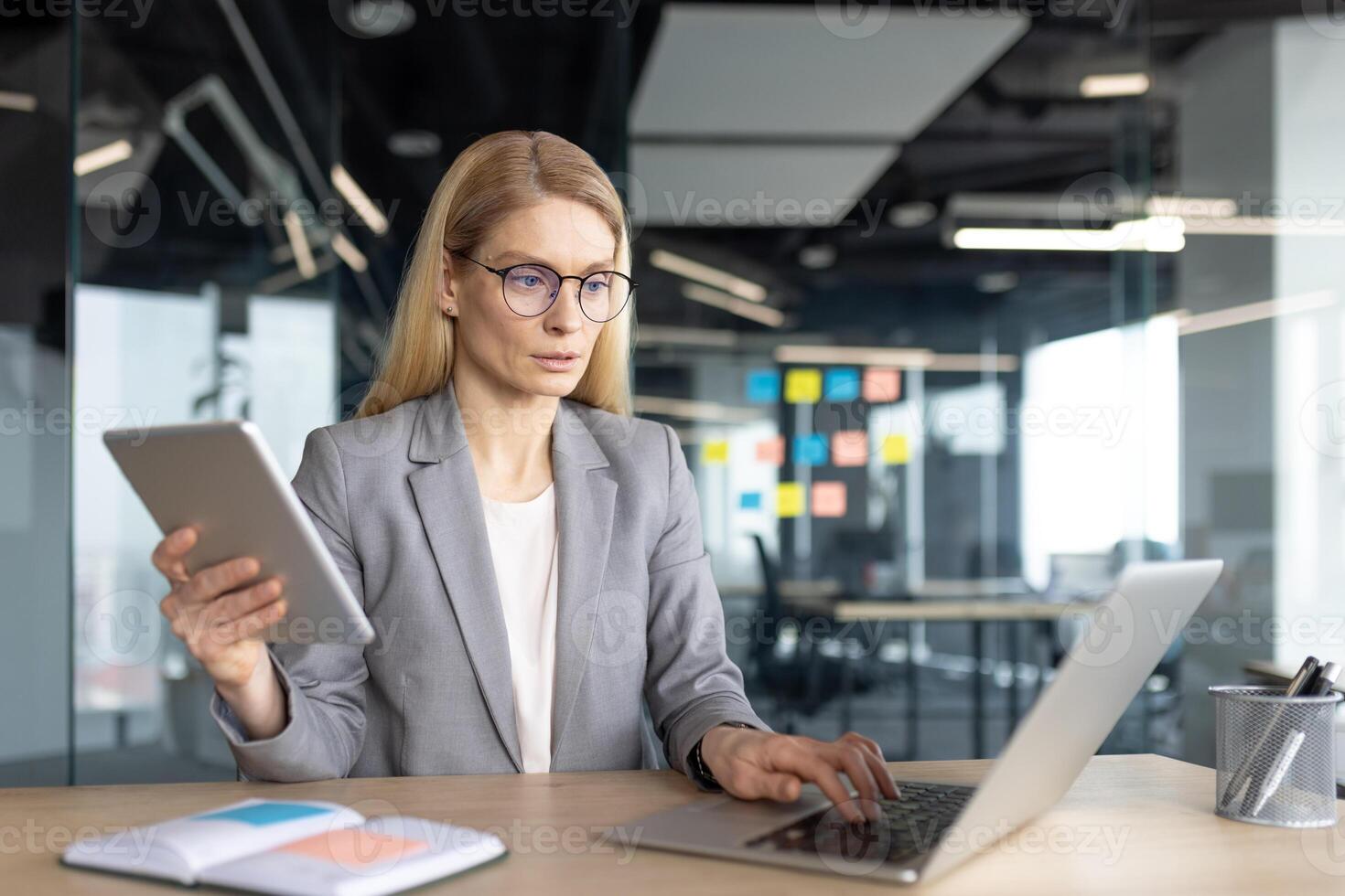A focused businesswoman working at a desk with a laptop and tablet in a modern office setting, considering work. photo