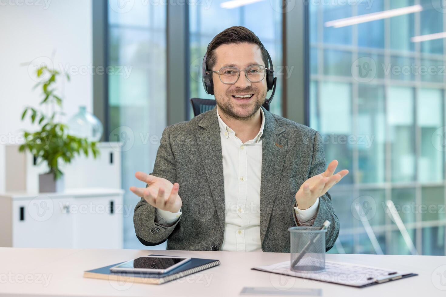 Portrait of a young smiling man sitting at a desk in an office, wearing a suit and headset, explaining and talking to the camera. photo