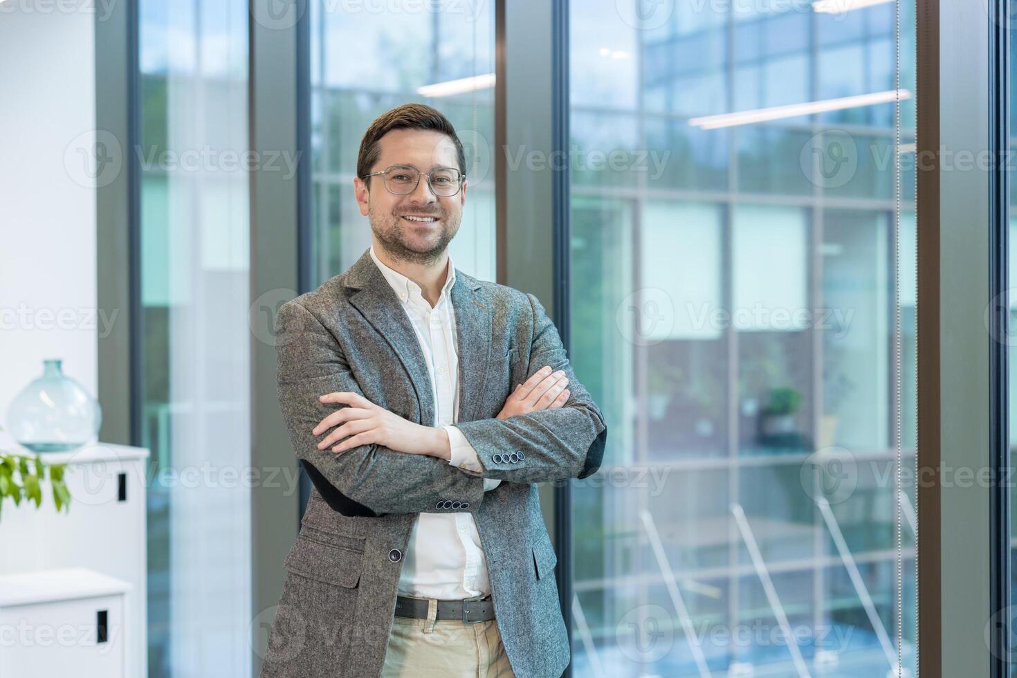 A smiling young man, founder and owner of a firm and company, stands in the lobby of an office building, arms crossed on his chest and confidently looking into the camera. photo