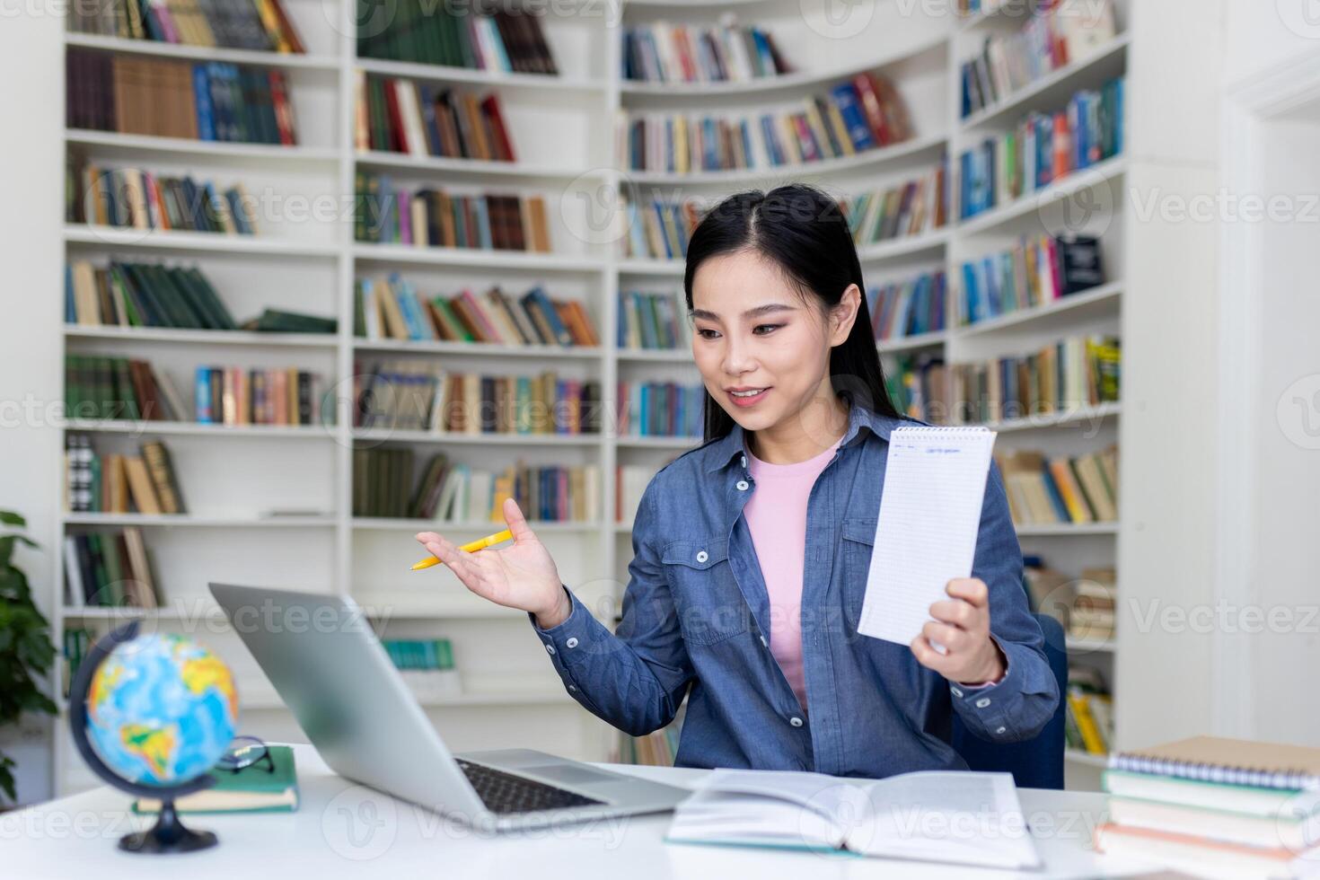 An Asian woman in a library setting, holding a notepad and giving a presentation online via her laptop. photo