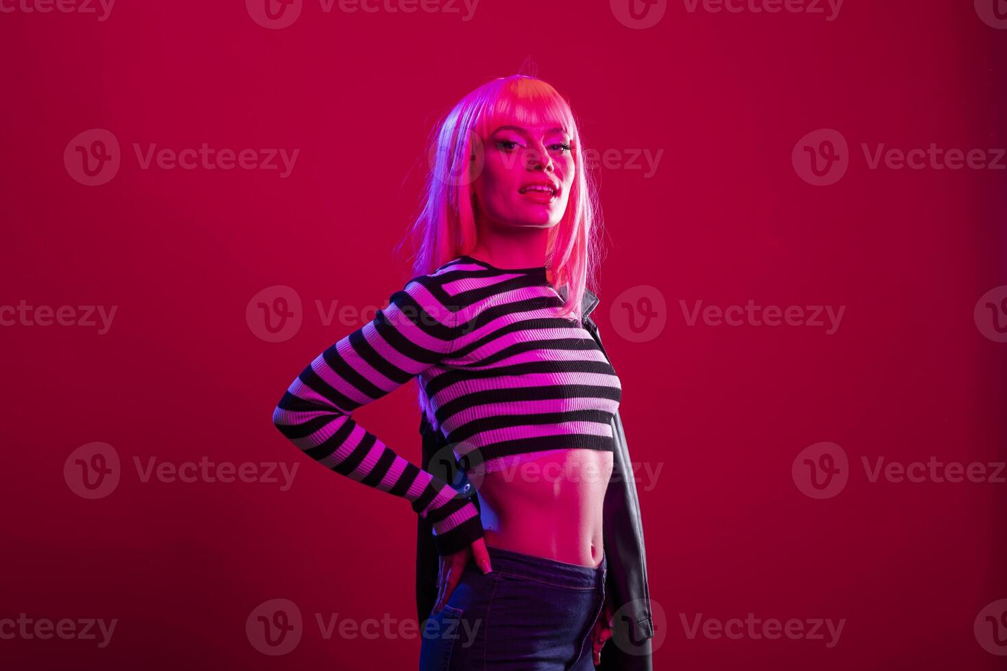 Female model with striped outfit and pink wig stands in calm yet assertive pose against isolated background, exuding vibrant energy. Glamorous lady with hand on waist, radiating confidence in studio. photo