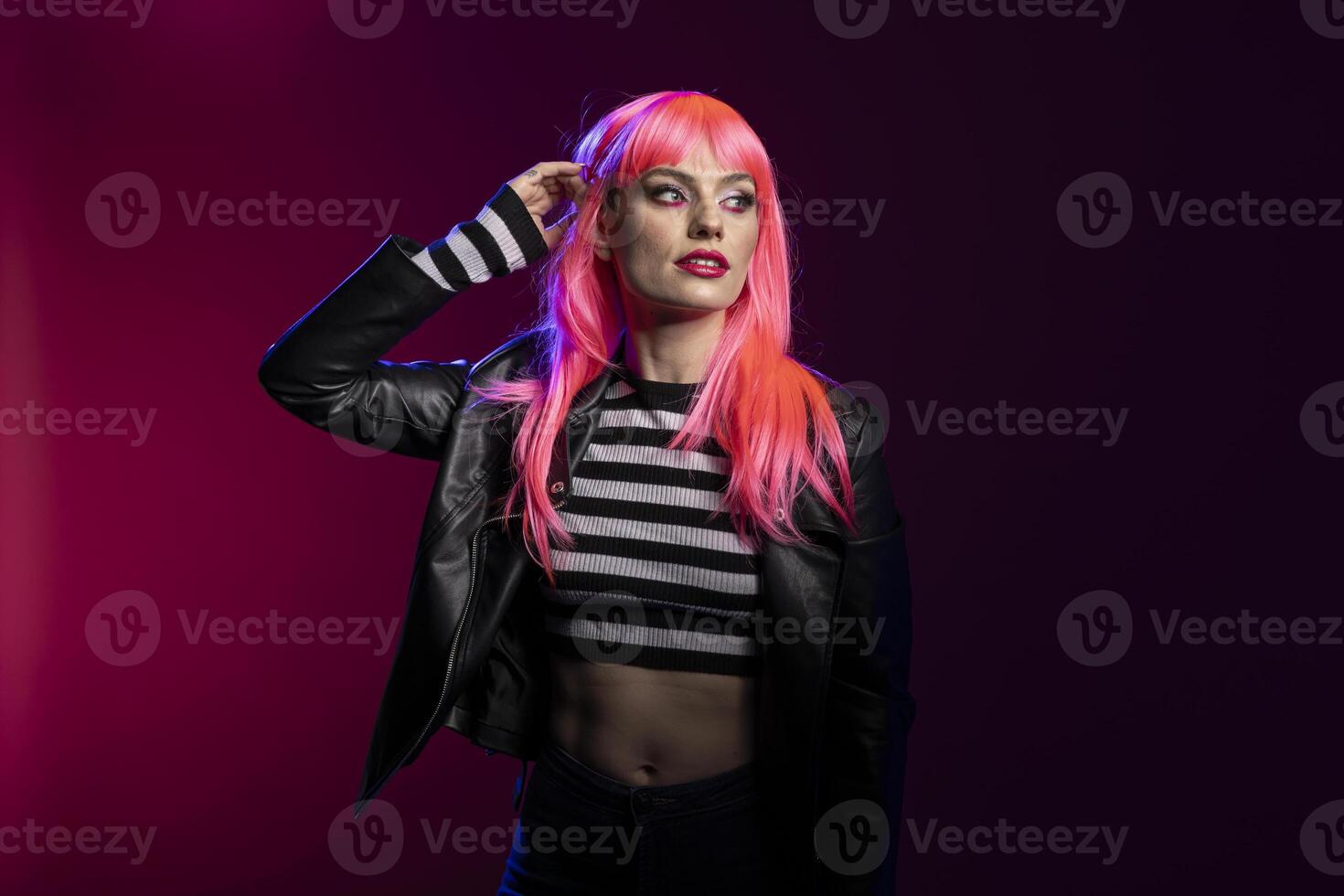 Young fashionable female with pink bangs hairstyle and eyeliner stands confidently with edgy outfit in studio portrait. Glamorous caucasian lady in leather jacket and crop top, posing with raised arm. photo