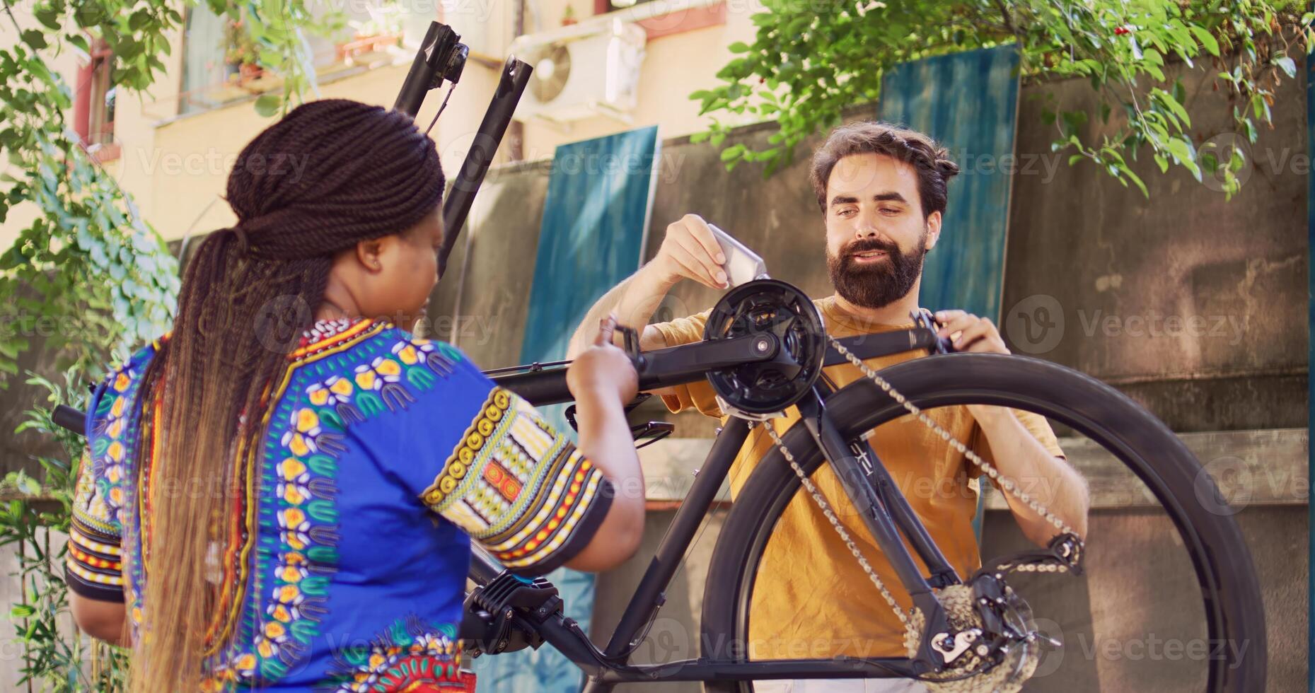 African american girlfriend assisting caucasian boyfriend with lubricating rusty bicycle chain outside as summer maintenance. Sporty multiethnic couple greasing bike chain in yard for safe cycling. photo